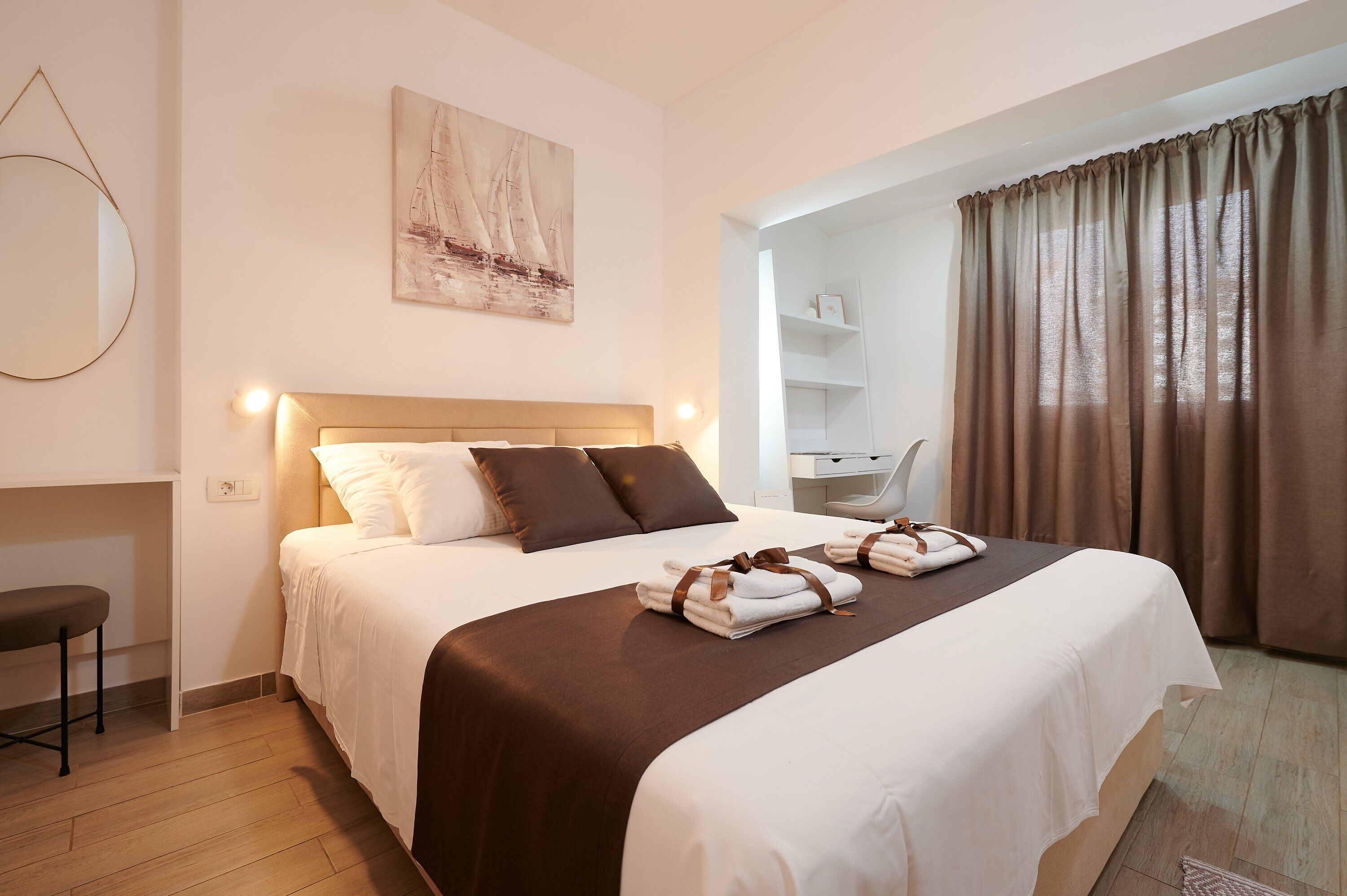 Una cama bien hecha con una colcha marrón y almohadas blancas, un espejo redondo en la pared y un cuadro sobre la cama.