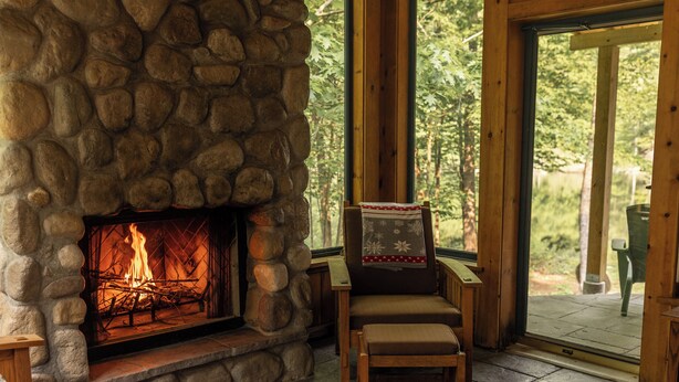 Une cheminée en pierre avec un feu visible, un fauteuil en bois avec un coussin, et une vue sur l’extérieur à travers une porte vitrée.
