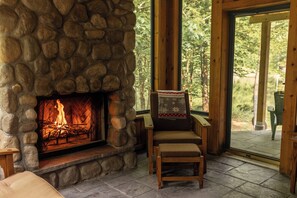 A stone fireplace with a visible fire, a wooden chair with a cushion, and a view of the outdoors through a glass door.