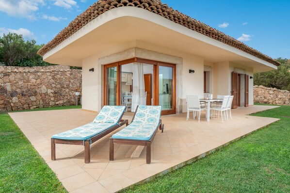 A house with a patio, two lounge chairs, a table with chairs, and a stone wall.