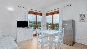 Een kamer met een bed, een televisie op een kast, een eettafel met stoelen, een koelkast en grote ramen met gordijnen.