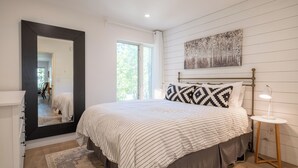 A bedroom with a large bed, a bedside table with a lamp, a mirror, and a window with a view of greenery. - Chic Condo | Queen Suite | Walk to Shops & Dining | Near Lake Tahoe (Kings Beach)