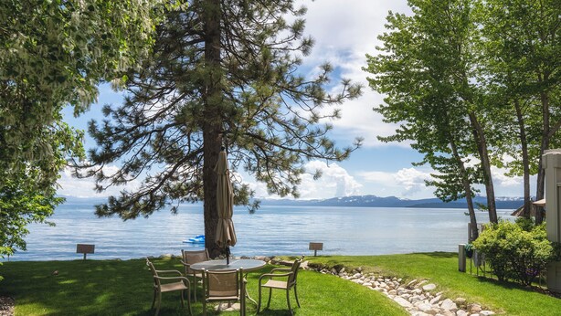 A lakeside patio with a table and chairs, surrounded by trees and a view of the lake. - Chic Condo | Queen Suite | Walk to Shops & Dining | Near Lake Tahoe (Kings Beach)