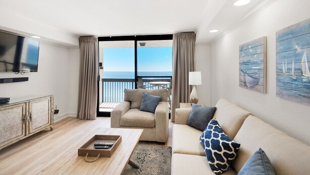 A modern living room with a sofa, armchair, and coffee table. There is a TV mounted on the wall, and a balcony with curtains.