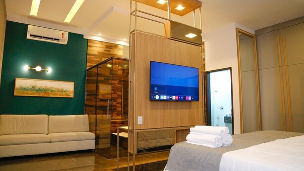 Una habitación de hotel moderna con un televisor de pantalla plana grande montado en un panel de madera, un sofá, una cama con toallas ordenadas y un separador de vidrio que divide la sala de estar del dormitorio.