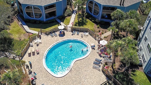 Un área de piscina con sillas de descanso y sombrillas, rodeada de edificios de forma octogonal y una piscina.