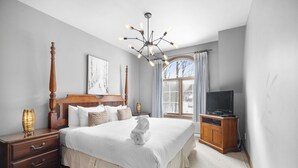 Une chambre avec un grand lit, une table de chevet en bois, un téléviseur posé sur une commode en bois et une fenêtre avec des rideaux.