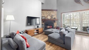 Un salon moderne avec des canapés gris, un téléviseur à écran plat, une cheminée en pierre et une grande fenêtre donnant sur la forêt.