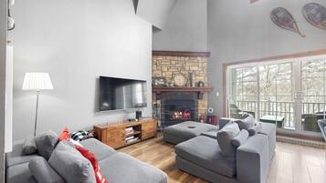 A modern living room with grey sofas, a flat-screen TV, a stone fireplace, and a large window offering a view of trees.