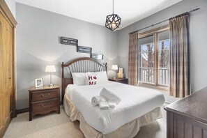 Un dormitorio ordenado con cama, mesitas de noche, una lámpara colgante y una ventana con cortinas.