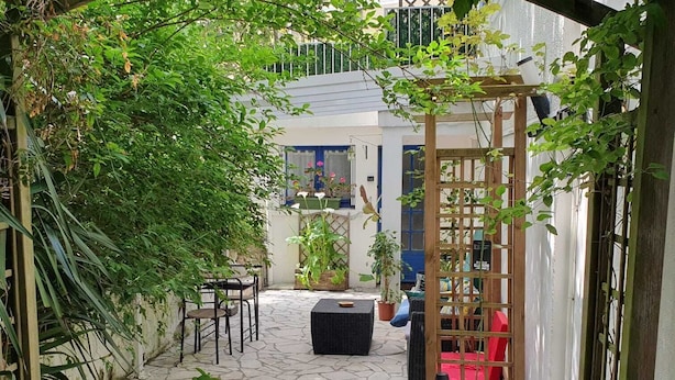 Een smal, met stenen bestraat binnenhof met een houten pergola begroeid met groen, een zwarte rieten bank en een rode stoel.