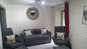 Een woonkamer met een grijze bank, twee fauteuils en een ronde spiegel aan de muur.