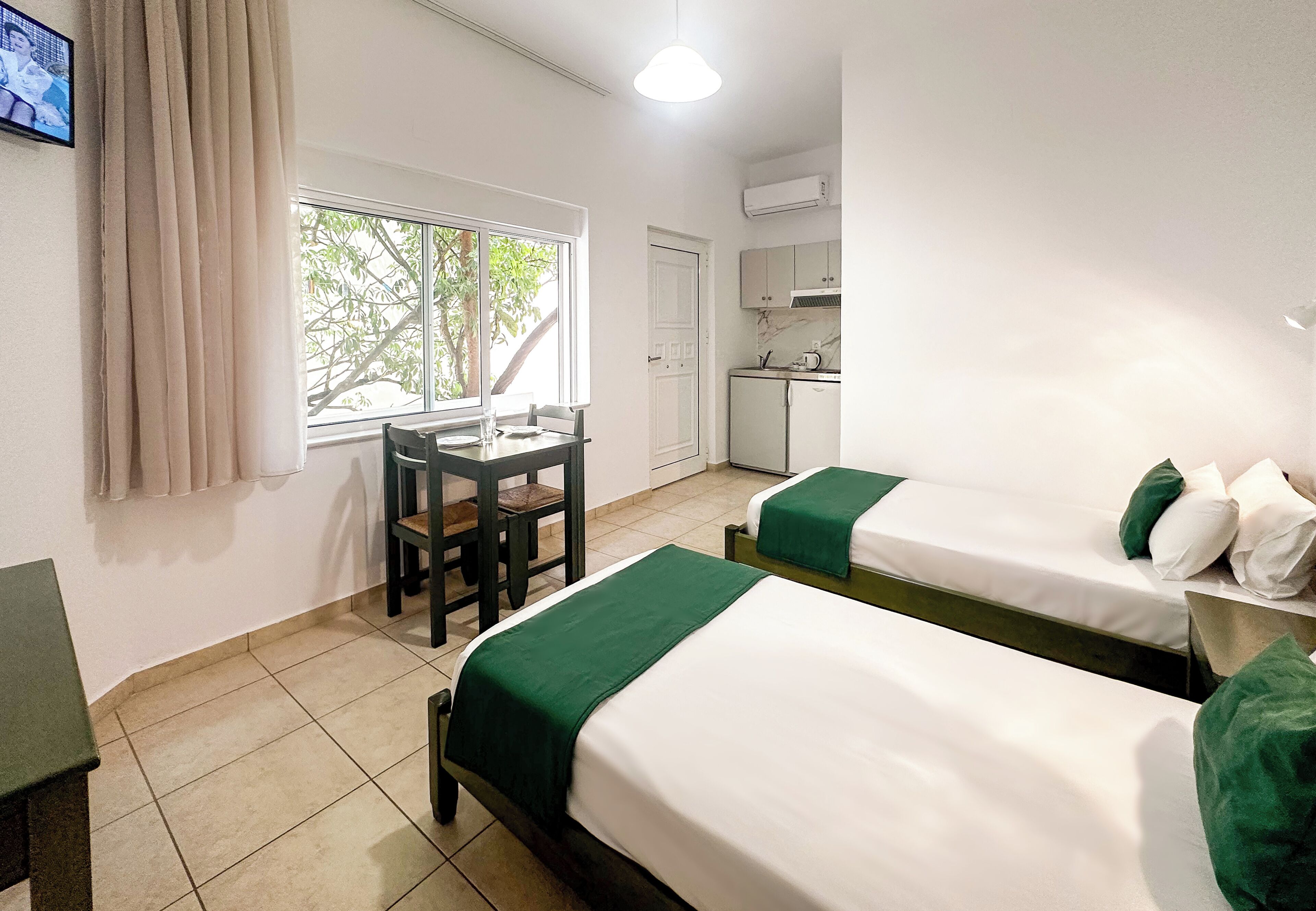 A hotel room with two beds, a small table, a chair, a window with a view of trees, and a kitchenette.