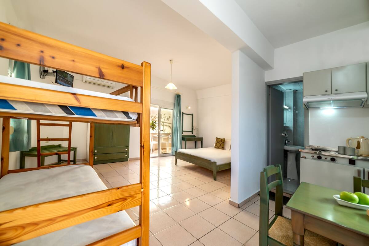 Una habitación compacta con literas, una cama individual, una mesa de comedor con sillas, una zona de cocina y un pequeño rincón de descanso con mesa y sillas.