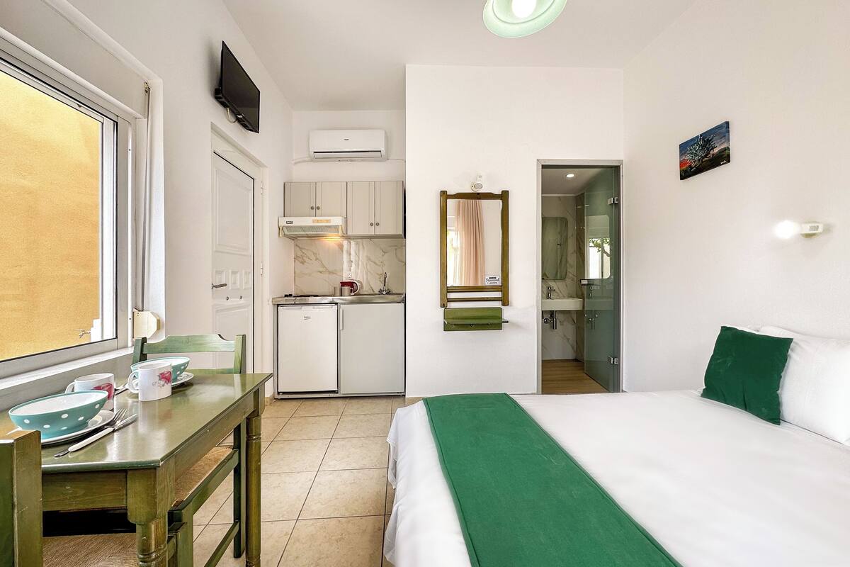 Habitación de hotel compacta con cama, kitchenette pequeña, mesa de comedor y televisor.
