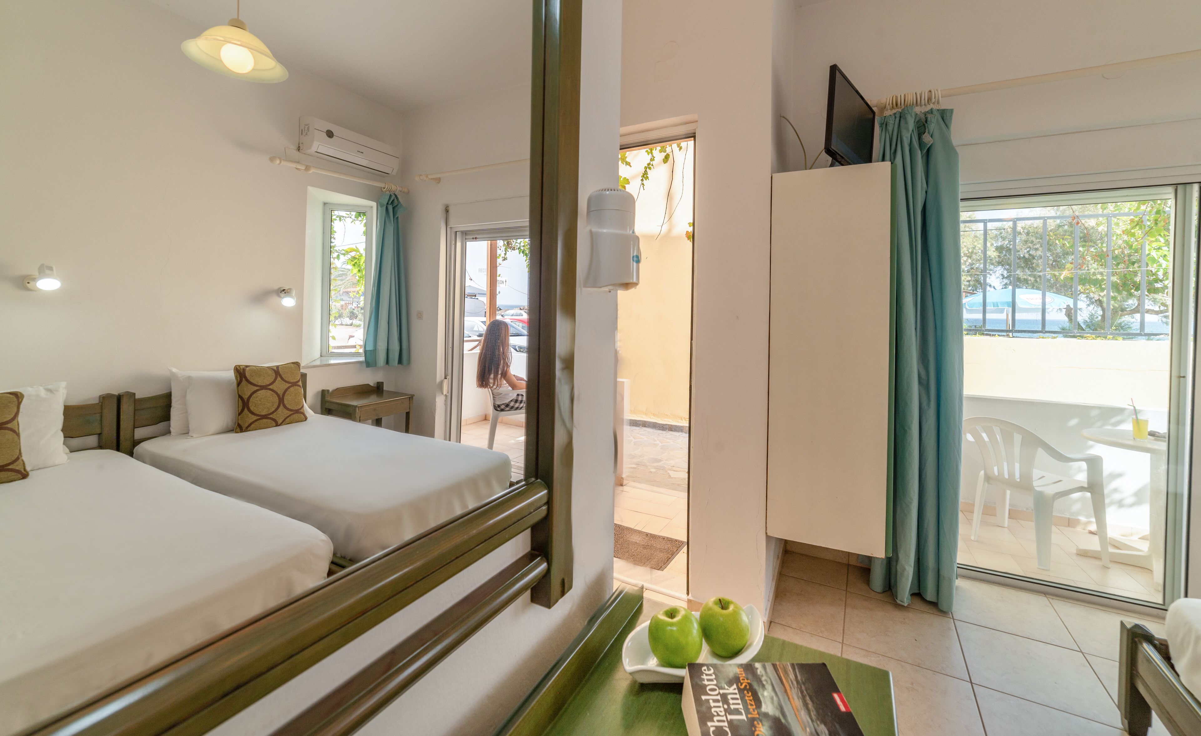 Une chambre d’hôtel avec deux lits, une petite table avec des pommes et un livre, et une vue sur l’extérieur.