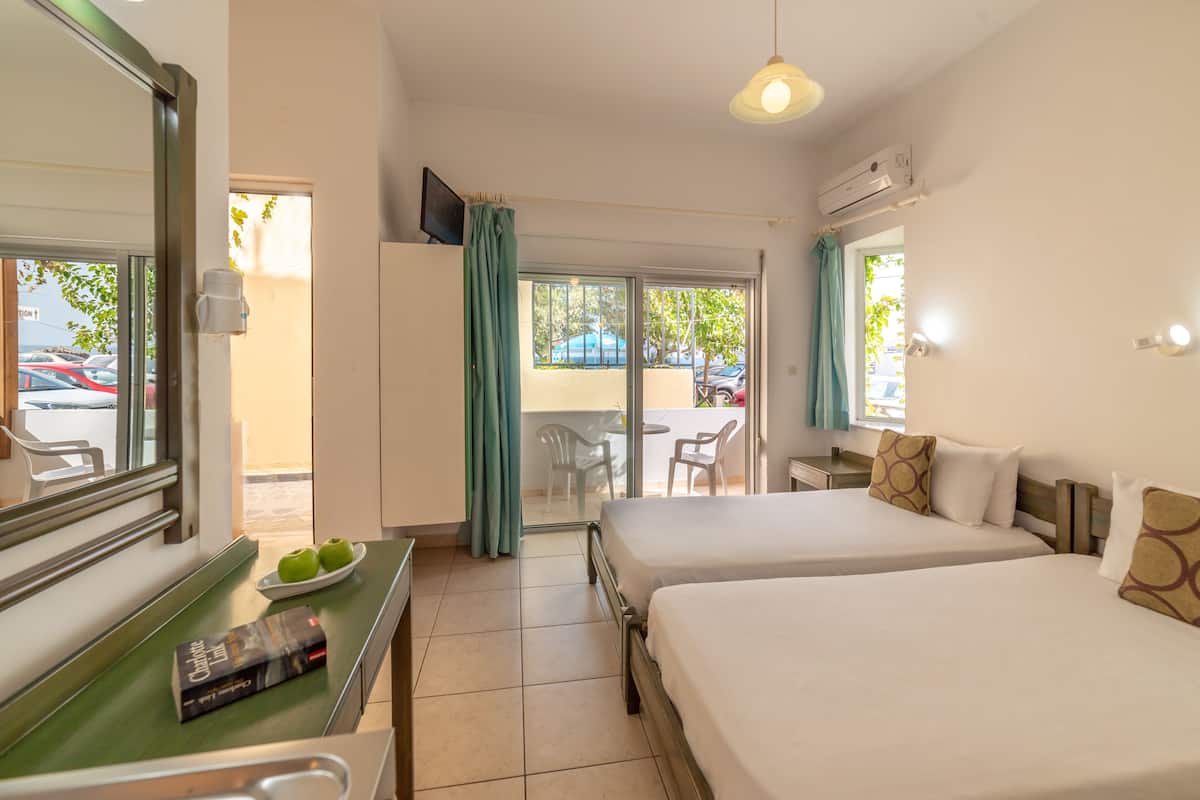Habitación de hotel con dos camas, una mesita con un libro y manzanas, una ventana con cortinas y un balcón con sillas y una mesa.