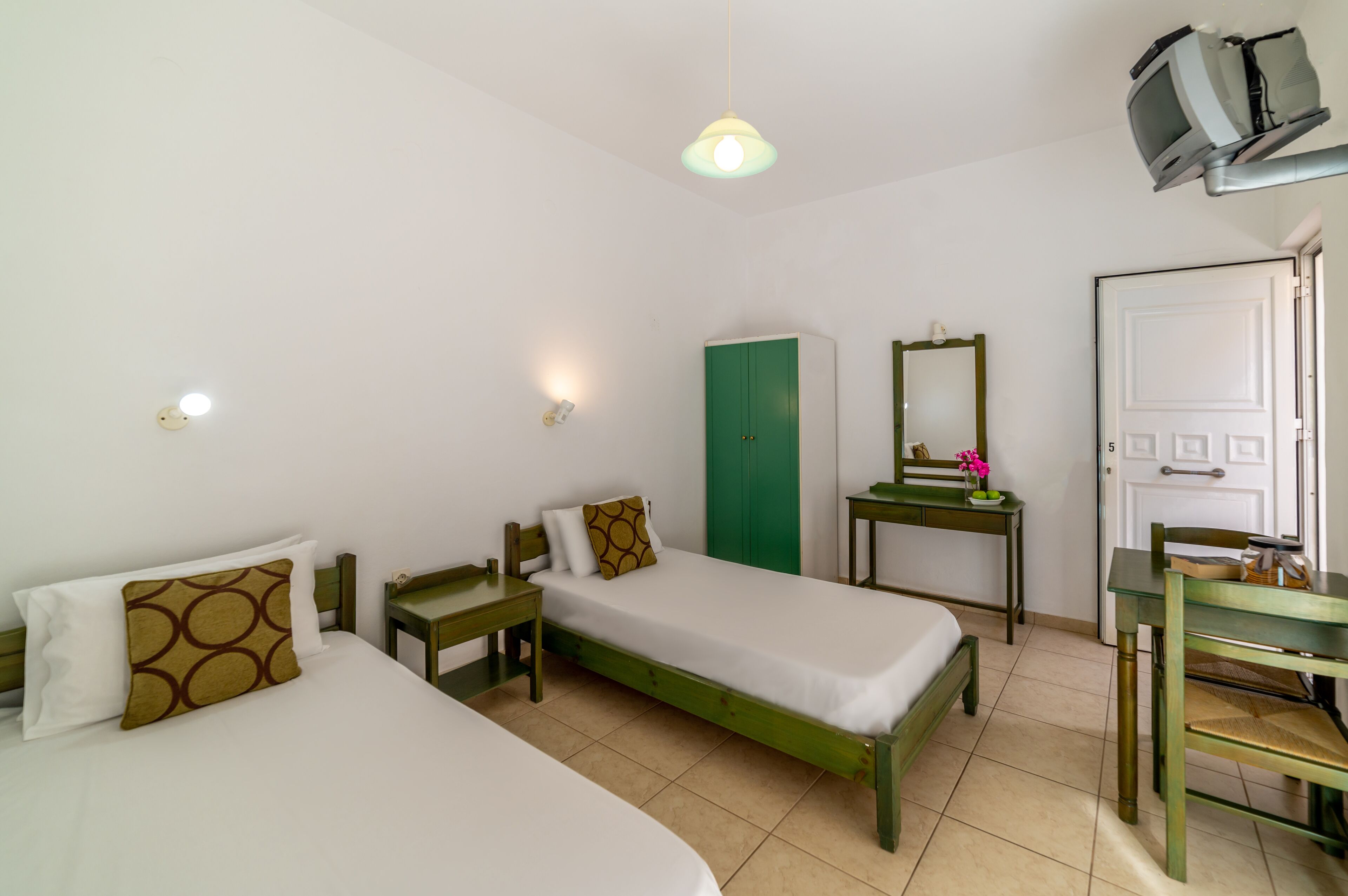 Una habitación pequeña y limpia con dos camas individuales, un armario verde, un escritorio con espejo y un televisor montado en la pared.