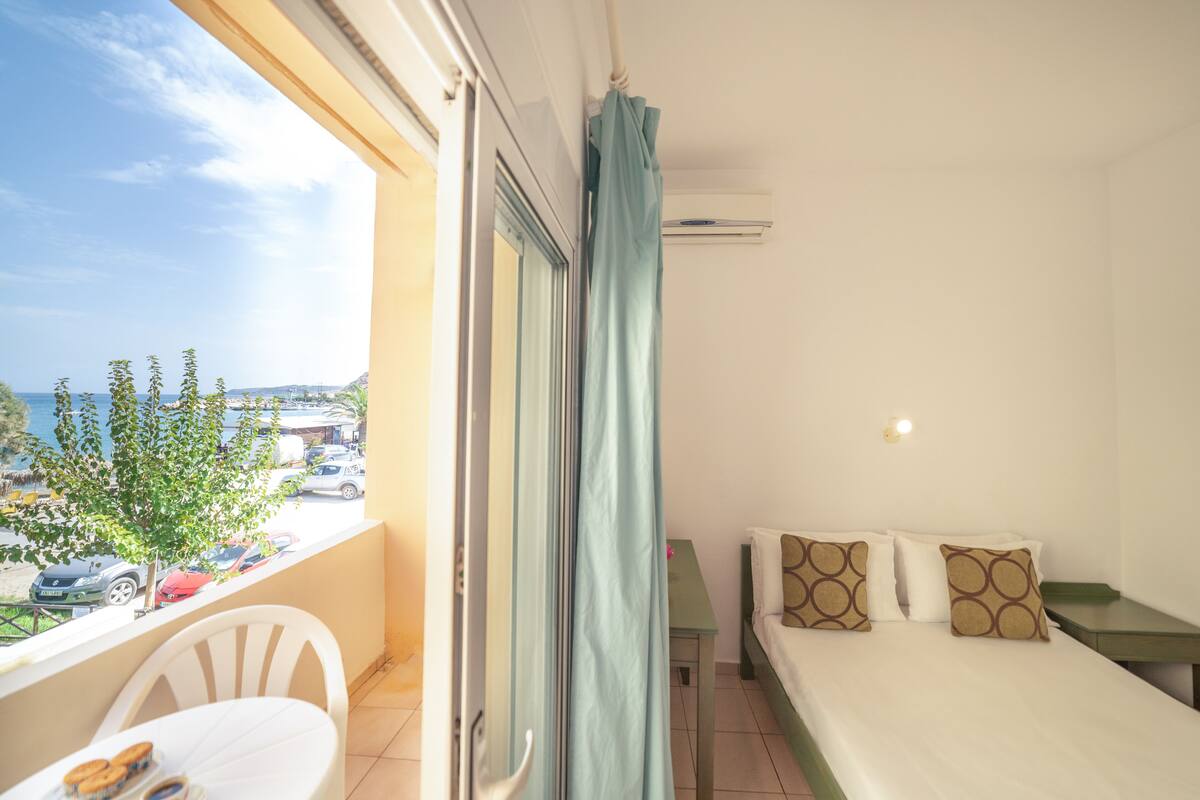 Un balcón con vista a la costa, una cama con almohadones estampados y una pequeña mesa con una silla.