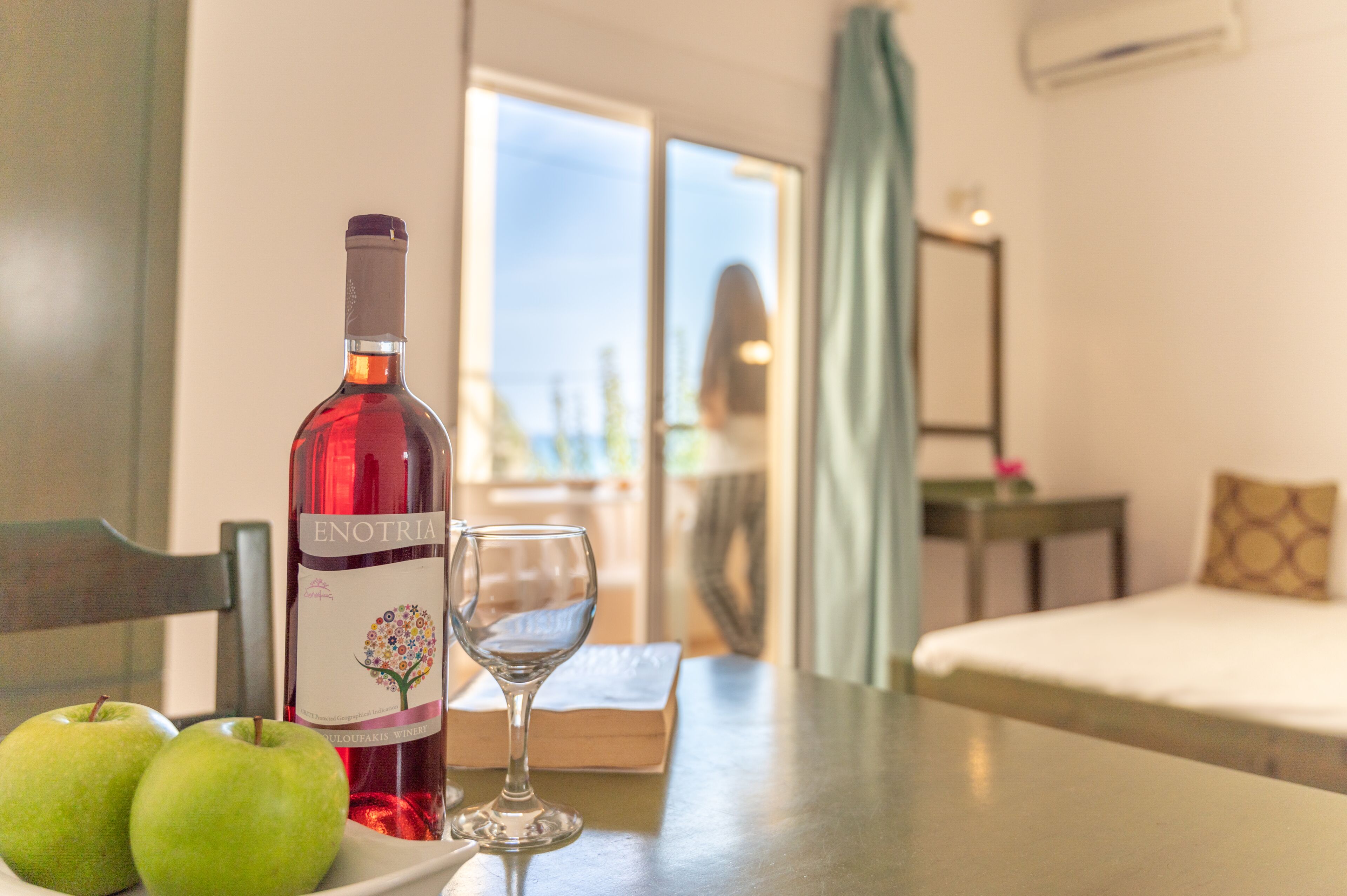 A hotel room with a table, a bottle of wine, a glass, and two apples.