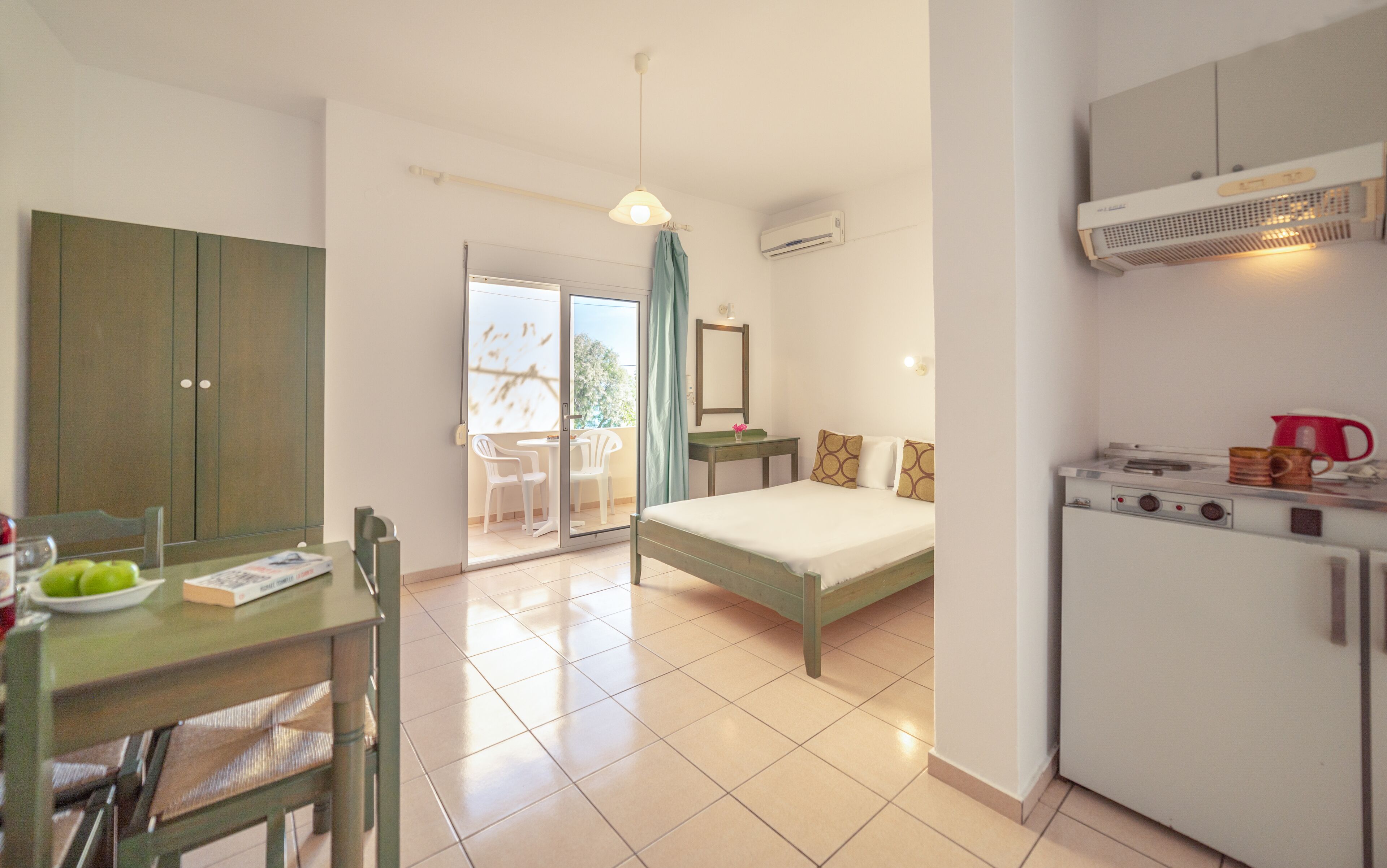 A compact hotel room with a bed, a small kitchenette, a dining area with a table and chairs, and a sliding glass door leading to an outdoor area.