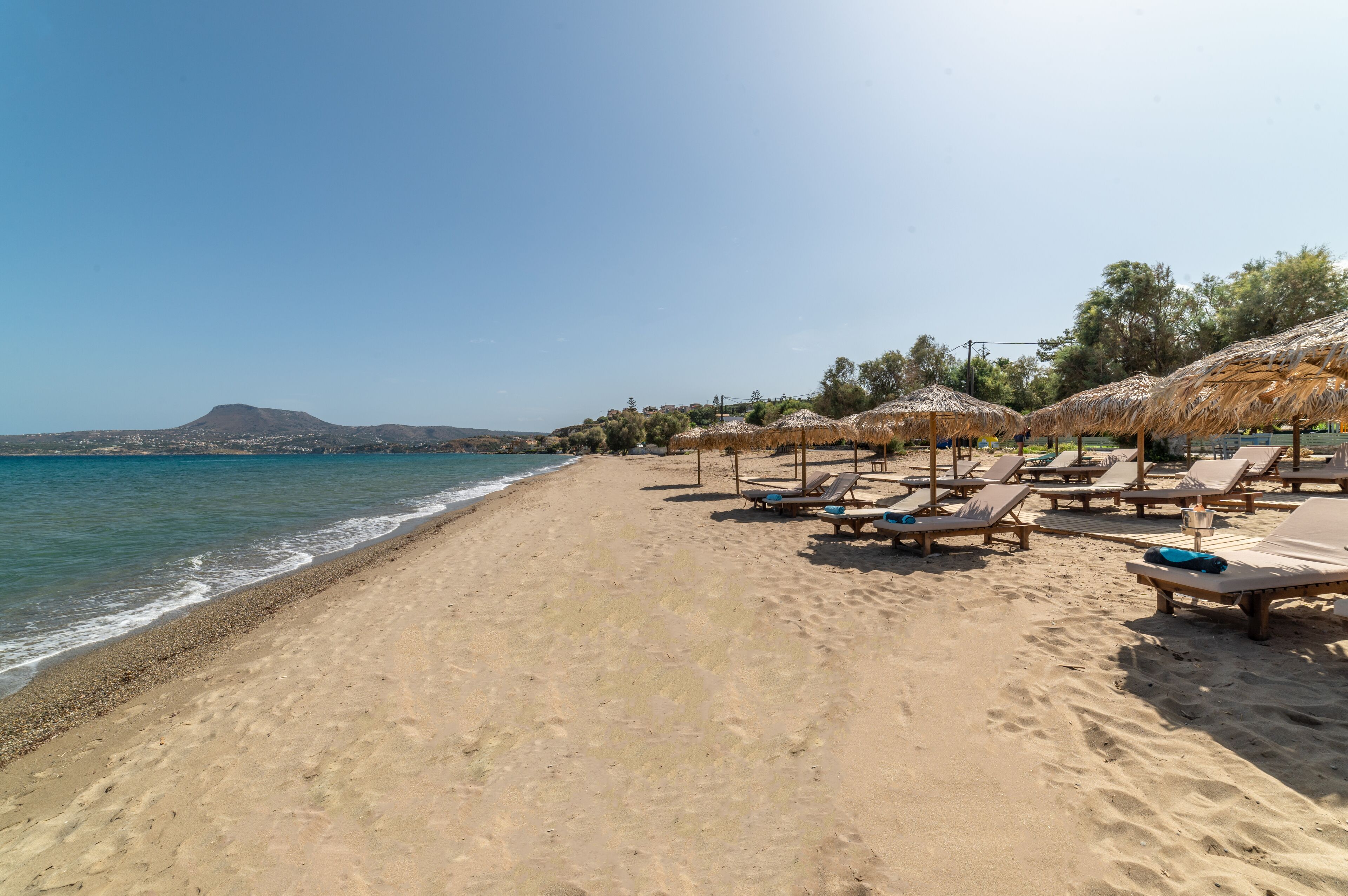 A beach with sun loungers and thatched umbrellas, a clear sky, and a distant mountain.