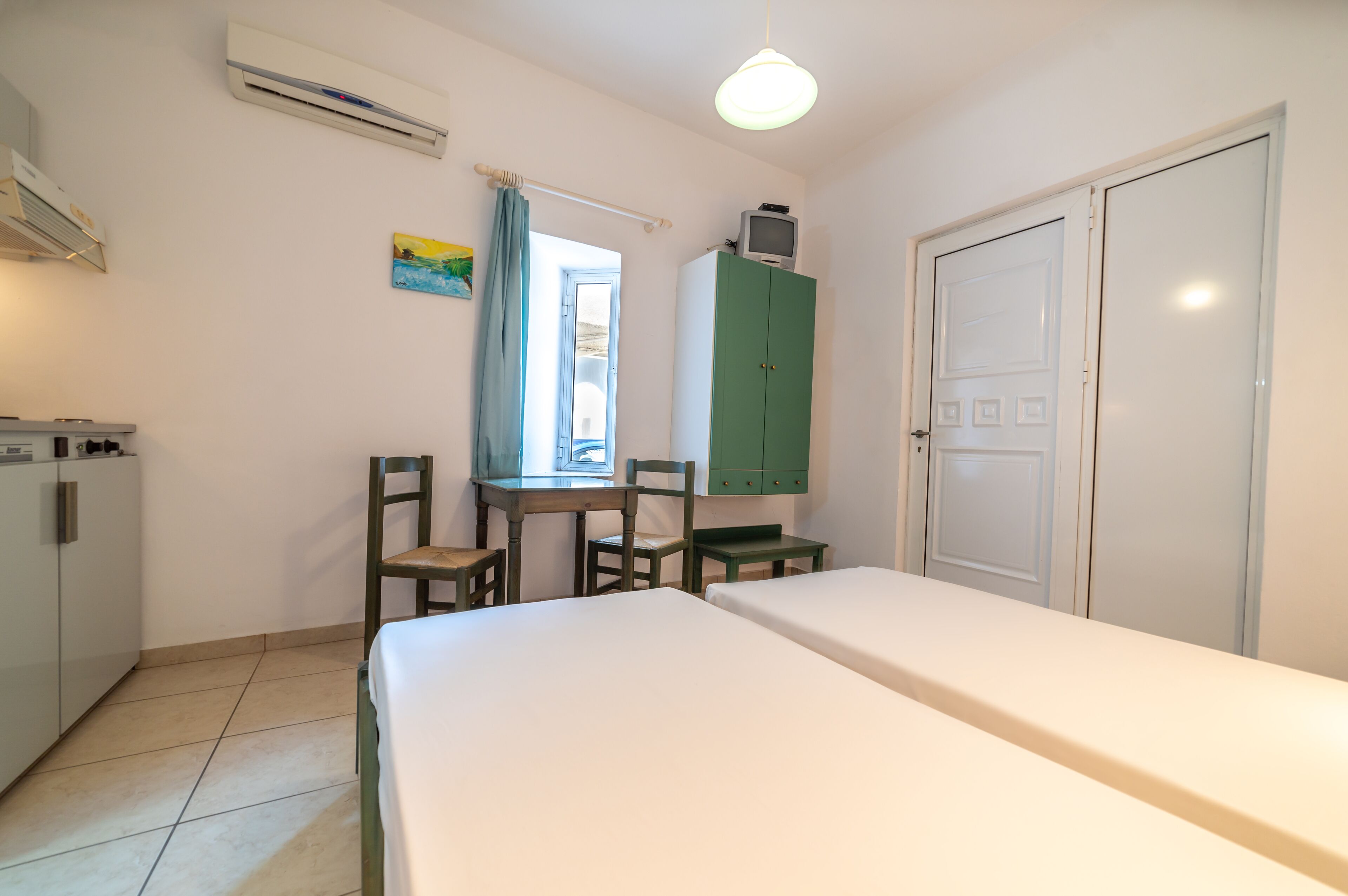 A room with a bed, a desk with chairs, a green cabinet, and a white door.