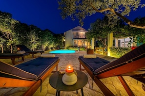 A poolside area with lounge chairs, a stone table with a candle, and a building with a pool in the background. - Villa Piano (Zupa dubrovacka)