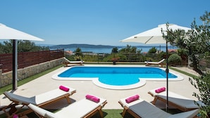 Une piscine entourée de chaises longues et d’un mur de pierre, agrémentée de parasols et offrant une vue sur la mer.