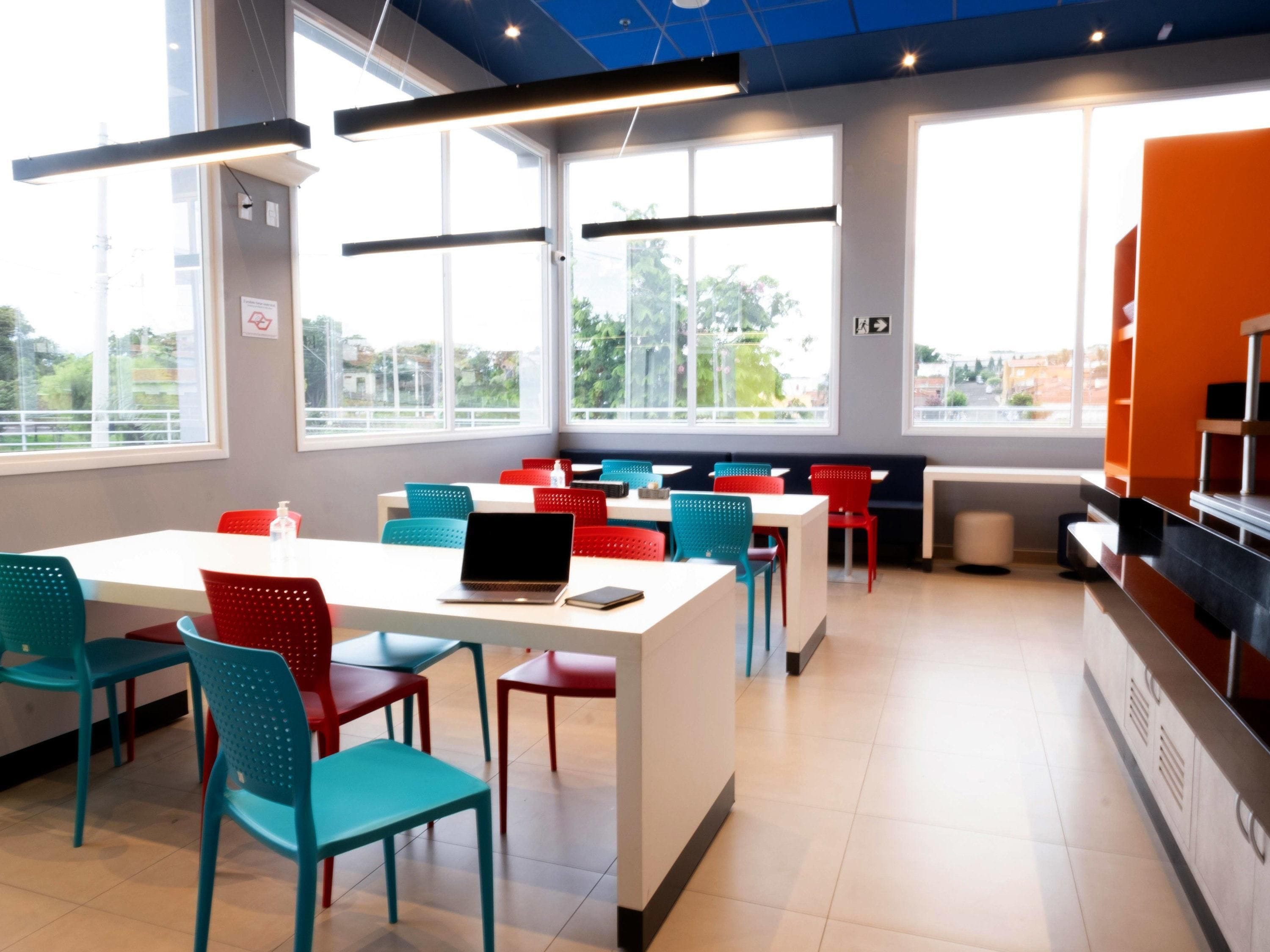 Un comedor moderno con mesas blancas y sillas de colores, ventanas amplias y un techo azul.