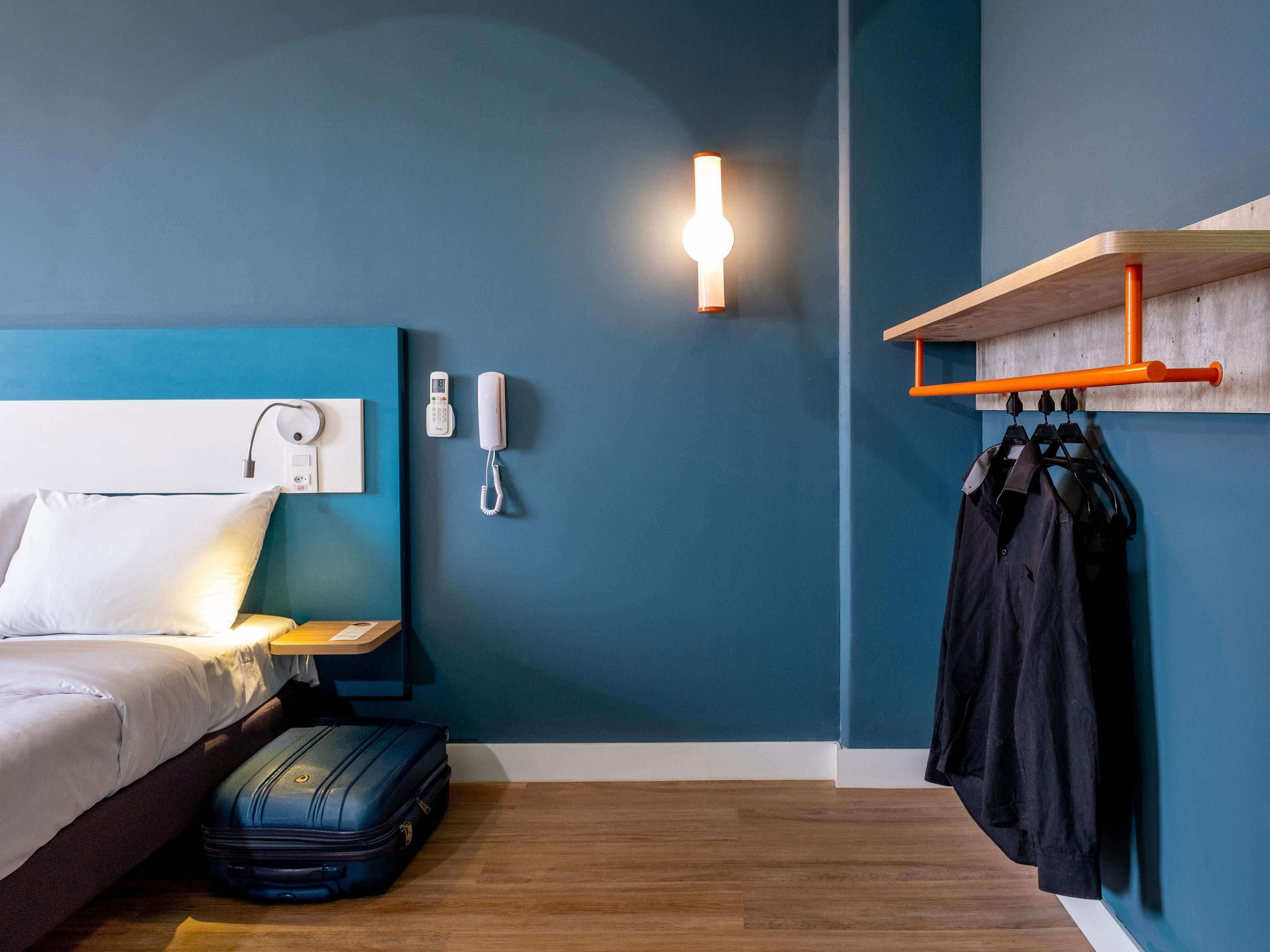 A hotel room with a bed, a bedside table, a lamp, a telephone, a suitcase, and a clothing rack.