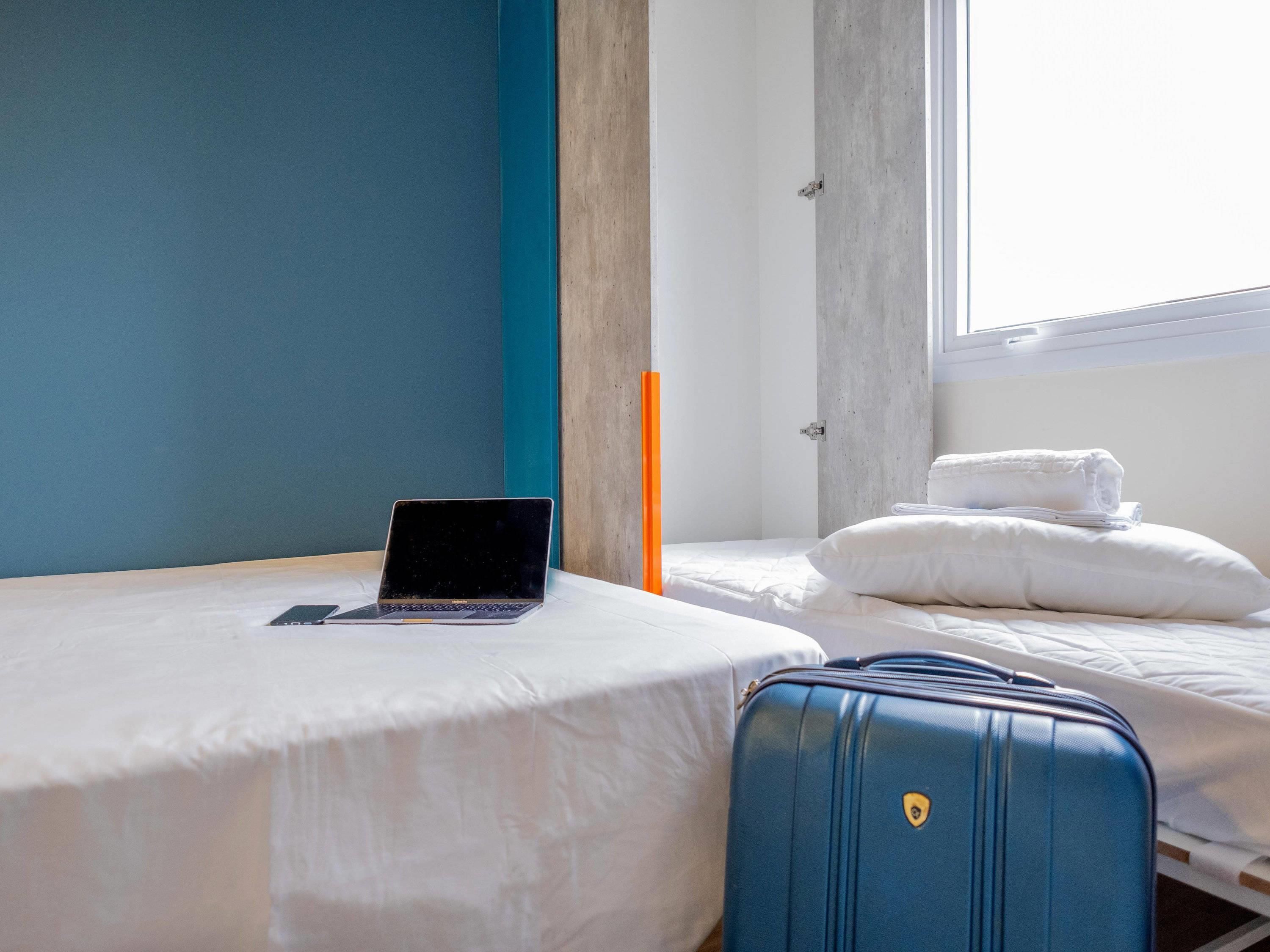 A hotel room with a bed, a laptop on the bed, a suitcase, and a window.