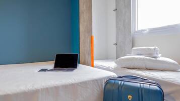 Una habitación de hotel con una cama, una computadora portátil sobre la cama, una maleta y una ventana.