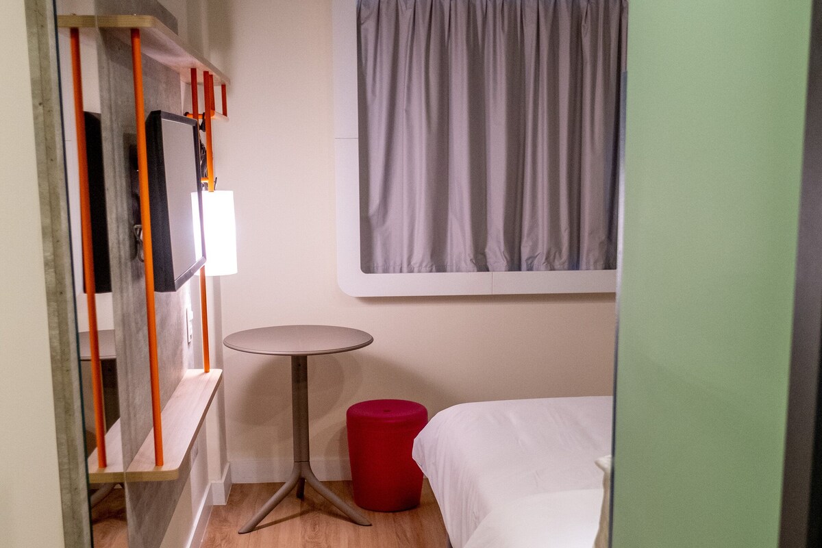 A small, compact room with a bed, a round table, a red stool, and a window with curtains.