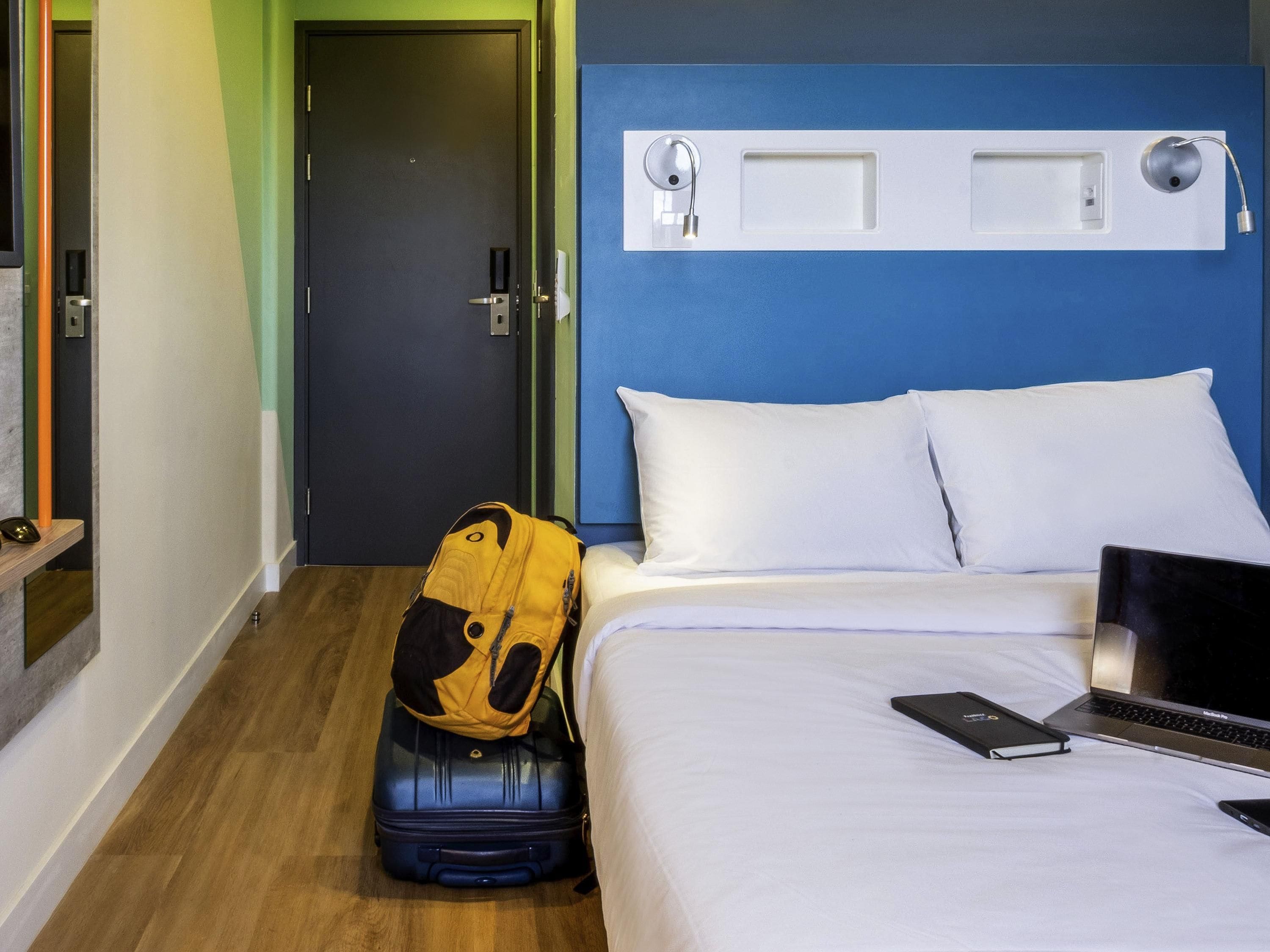 A hotel room with a bed, a suitcase, a laptop, and a backpack.