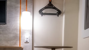 Una habitación de hotel con cama, una mesita redonda pequeña, un taburete rojo, una lámpara de pared y un perchero negro.