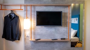 A modern hotel room with a mounted TV, a bedside table with a lamp, a mirror, and a hanging coat rack. - ibis budget Leme (Leme)