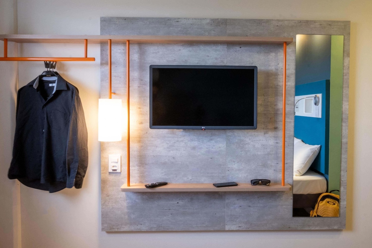 A modern hotel room with a mounted TV, a bedside table with a lamp, a mirror, and a hanging coat rack.