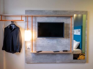 A modern hotel room with a mounted TV, a bedside table with a lamp, a mirror, and a hanging coat rack. - ibis budget Leme (Leme)