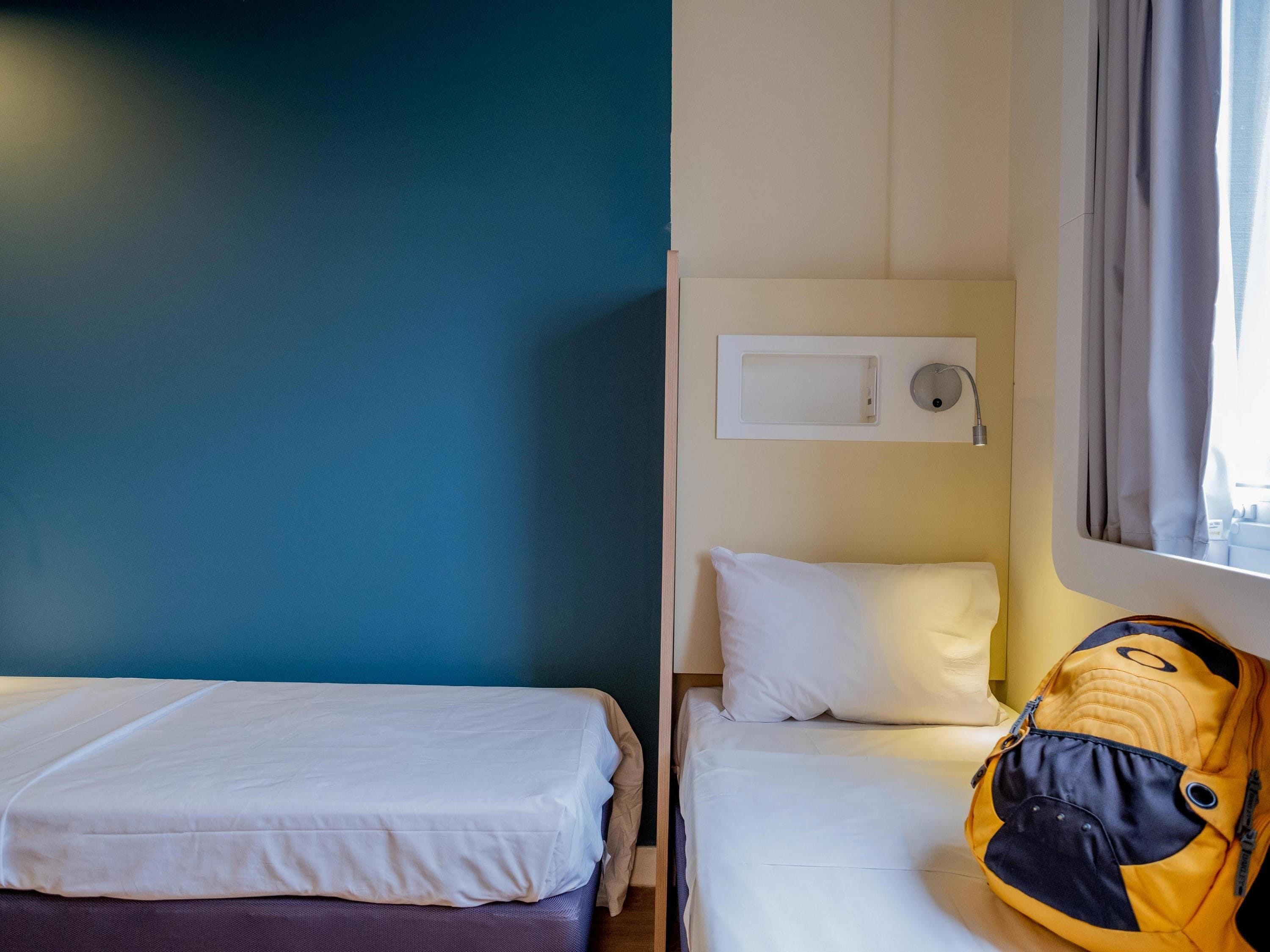 A hotel room with two beds, a window with curtains, a wall-mounted air conditioning unit, and a backpack on the floor.