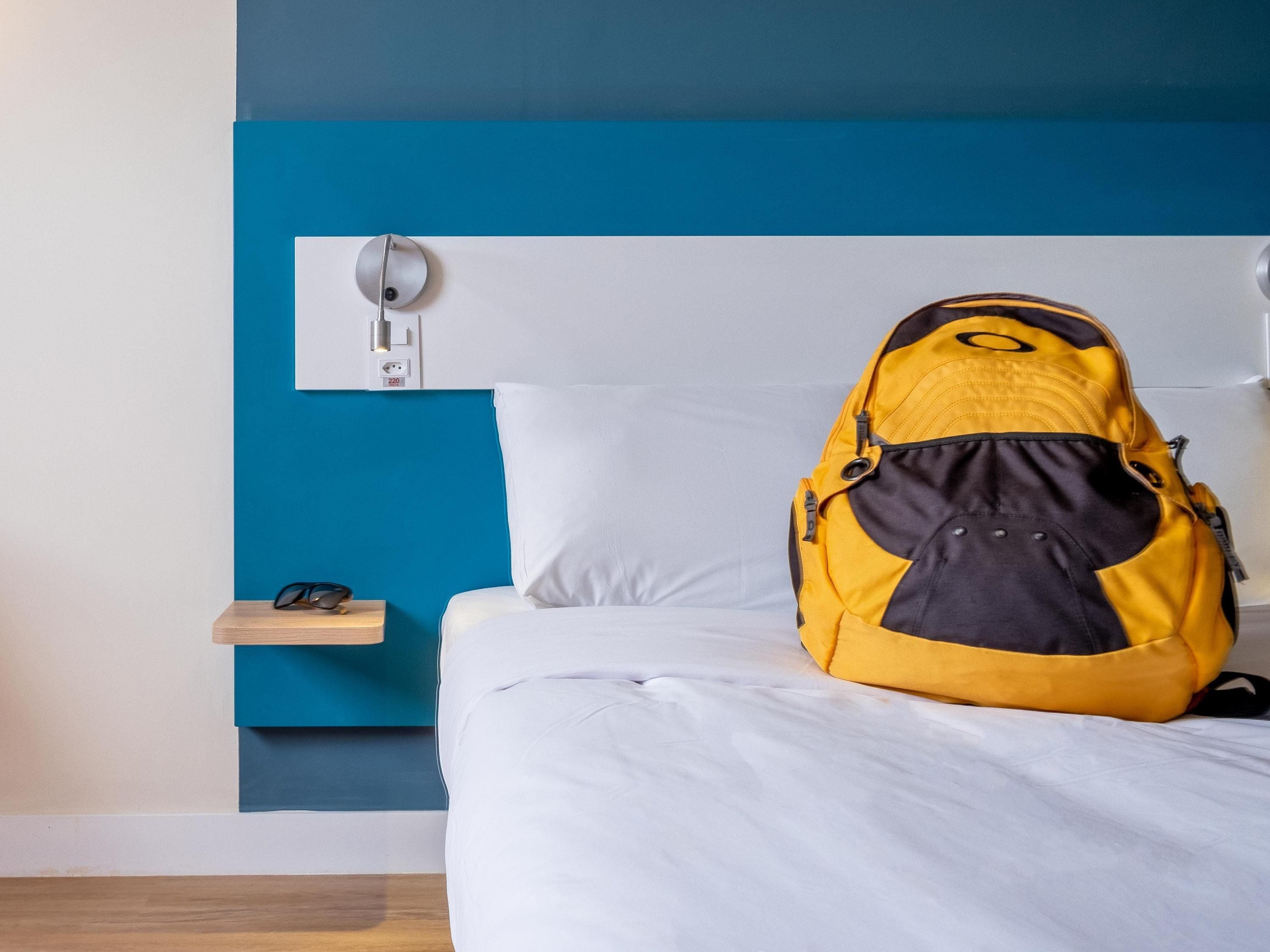 Una cama con ropa de cama blanca, una pared azul y una mochila negra y amarilla sobre la cama.