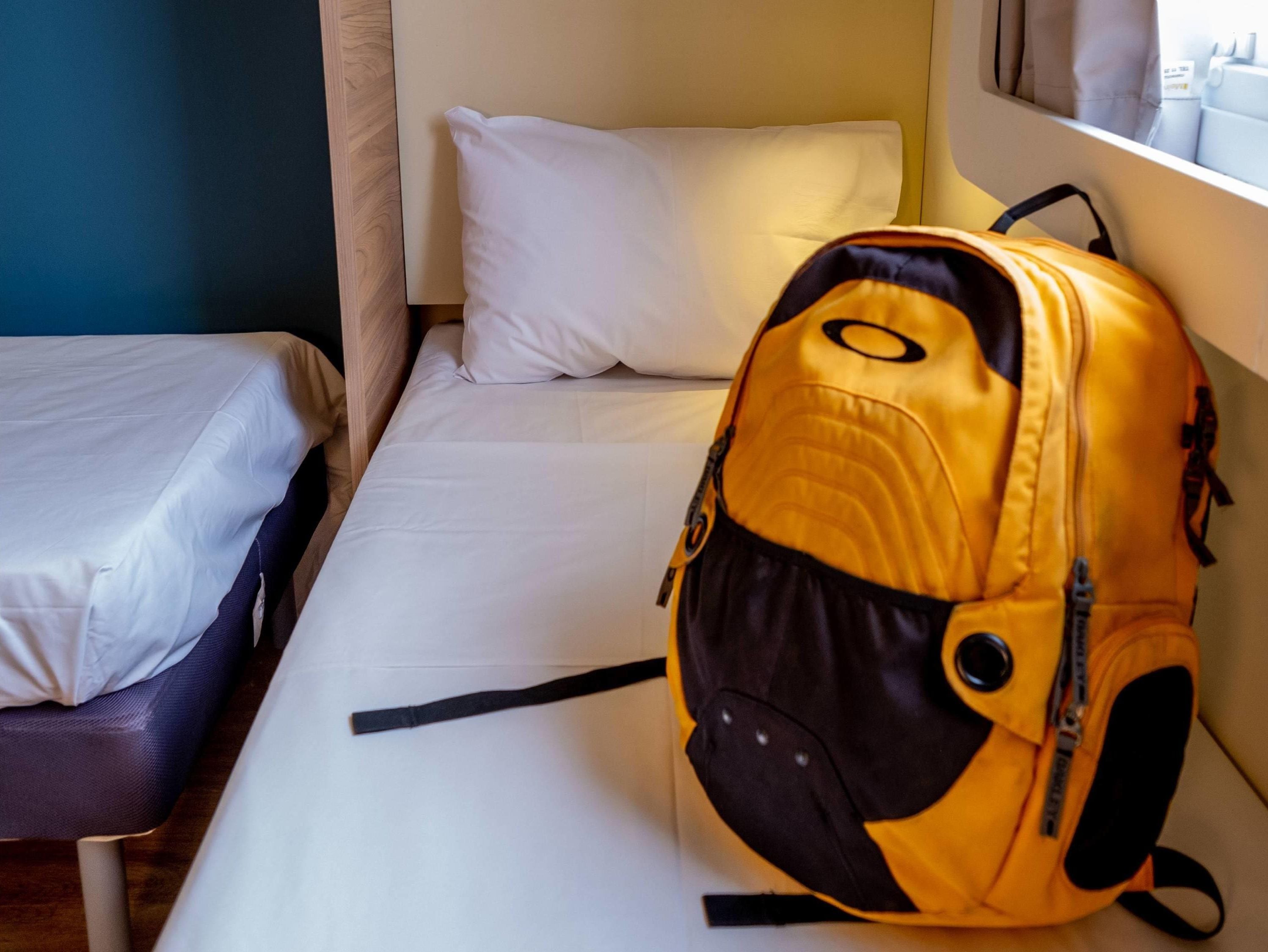 A single bed with white bedding, a small table, and a yellow backpack on the bed.