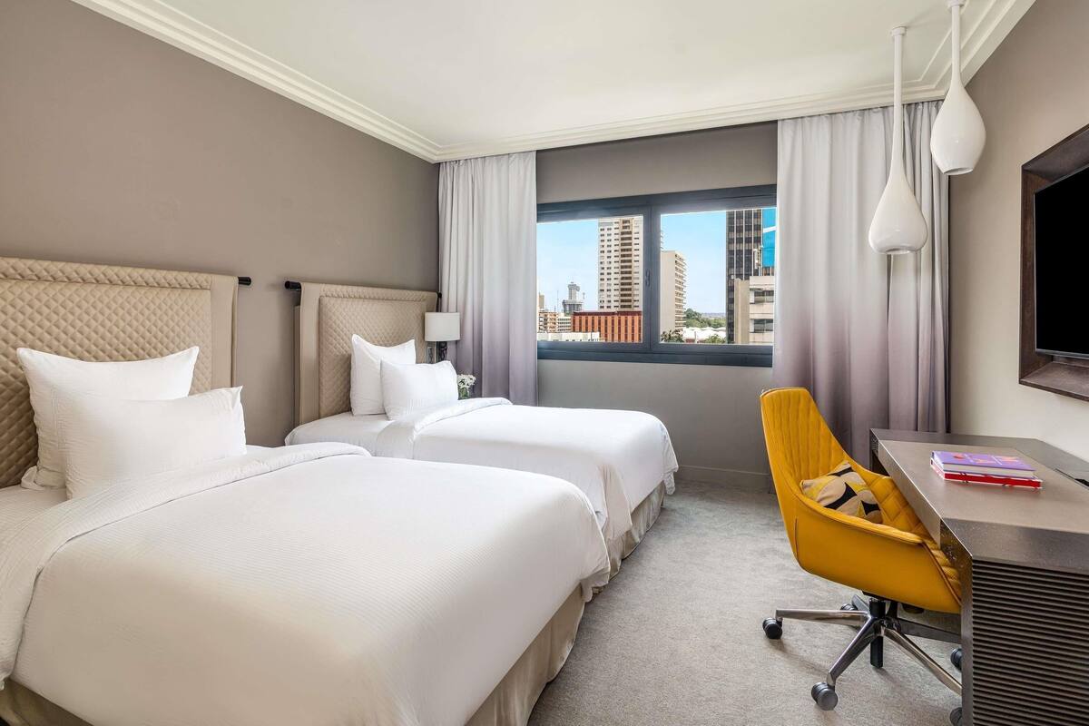 Ein Hotelzimmer mit zwei Betten, einem Schreibtisch, einem Stuhl, einem Fernseher und einem Fenster mit Blick auf die Stadt.