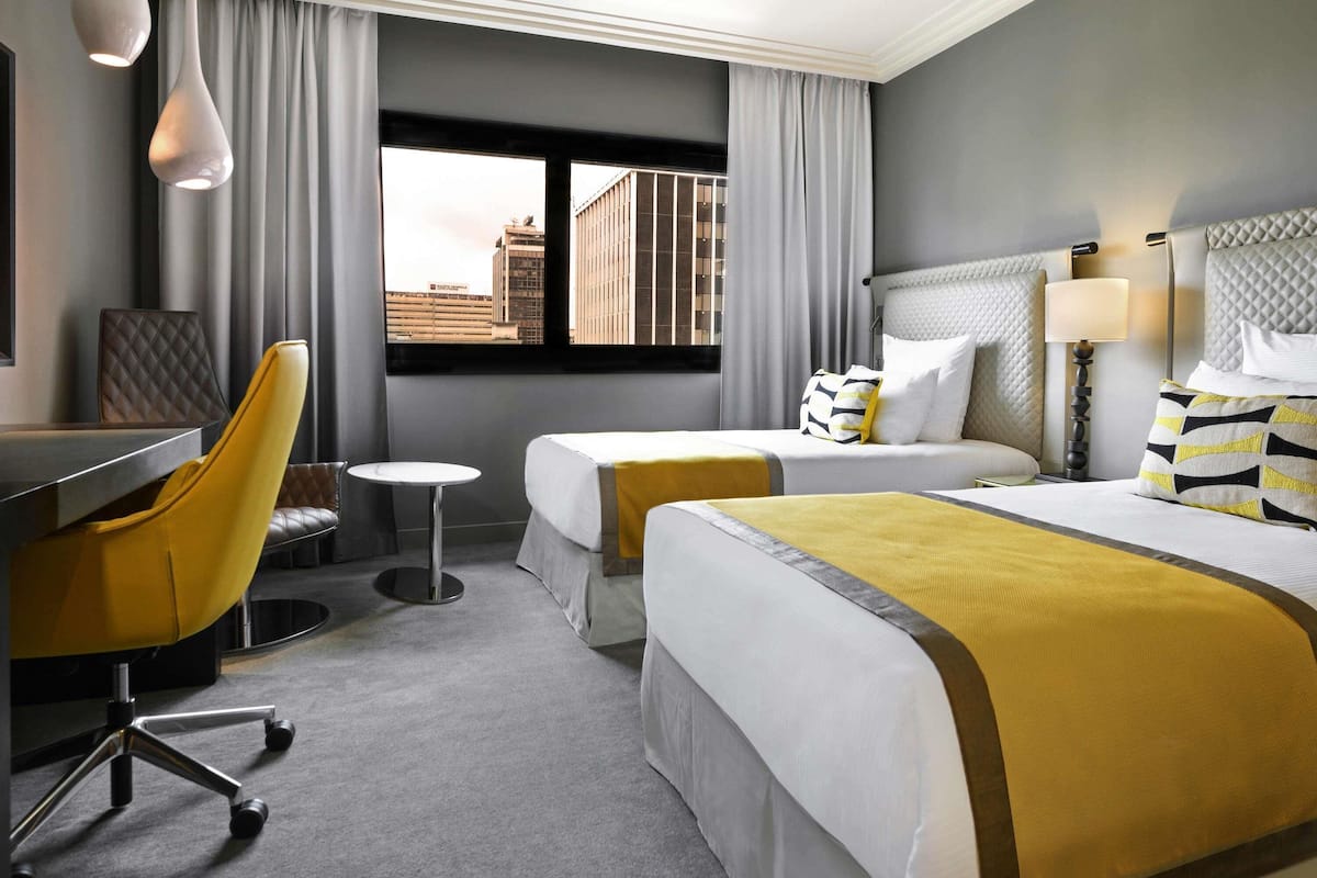 Ein Hotelzimmer mit zwei Betten, einem Schreibtisch, einem Stuhl und einem Fenster mit Blick auf die Stadt.