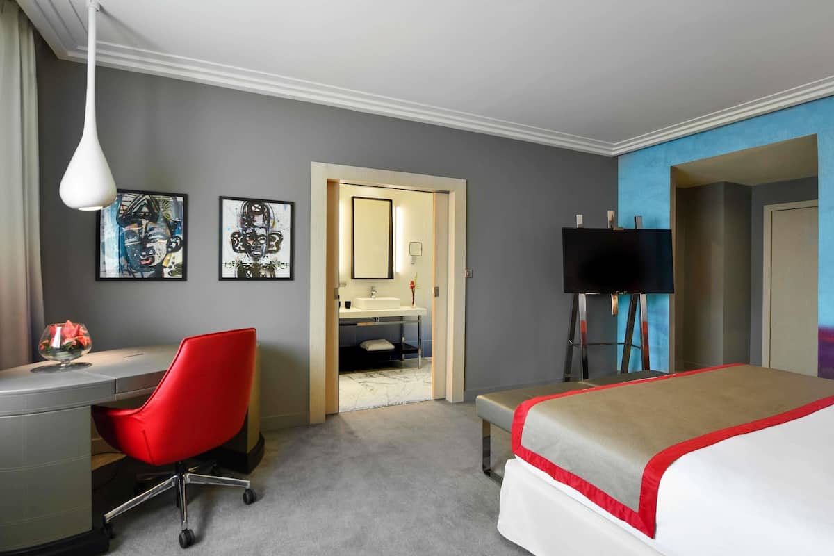 Ein Hotelzimmer mit Schreibtisch, rotem Sessel, Bett, Kunstwerk und Fernseher.
