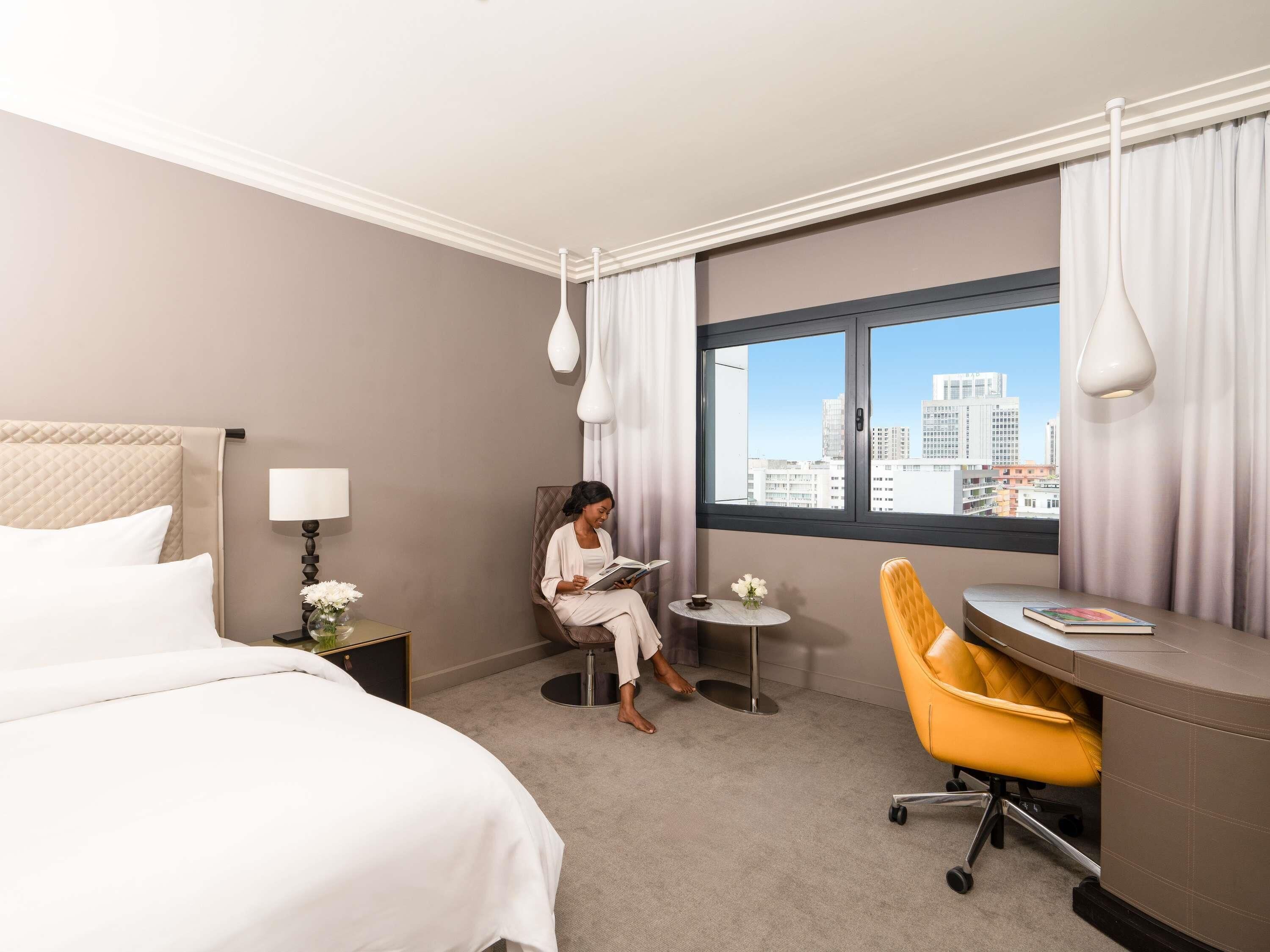 A hotel room with a bed, a desk, a chair, a lamp, a vase with flowers, and a view of the city.