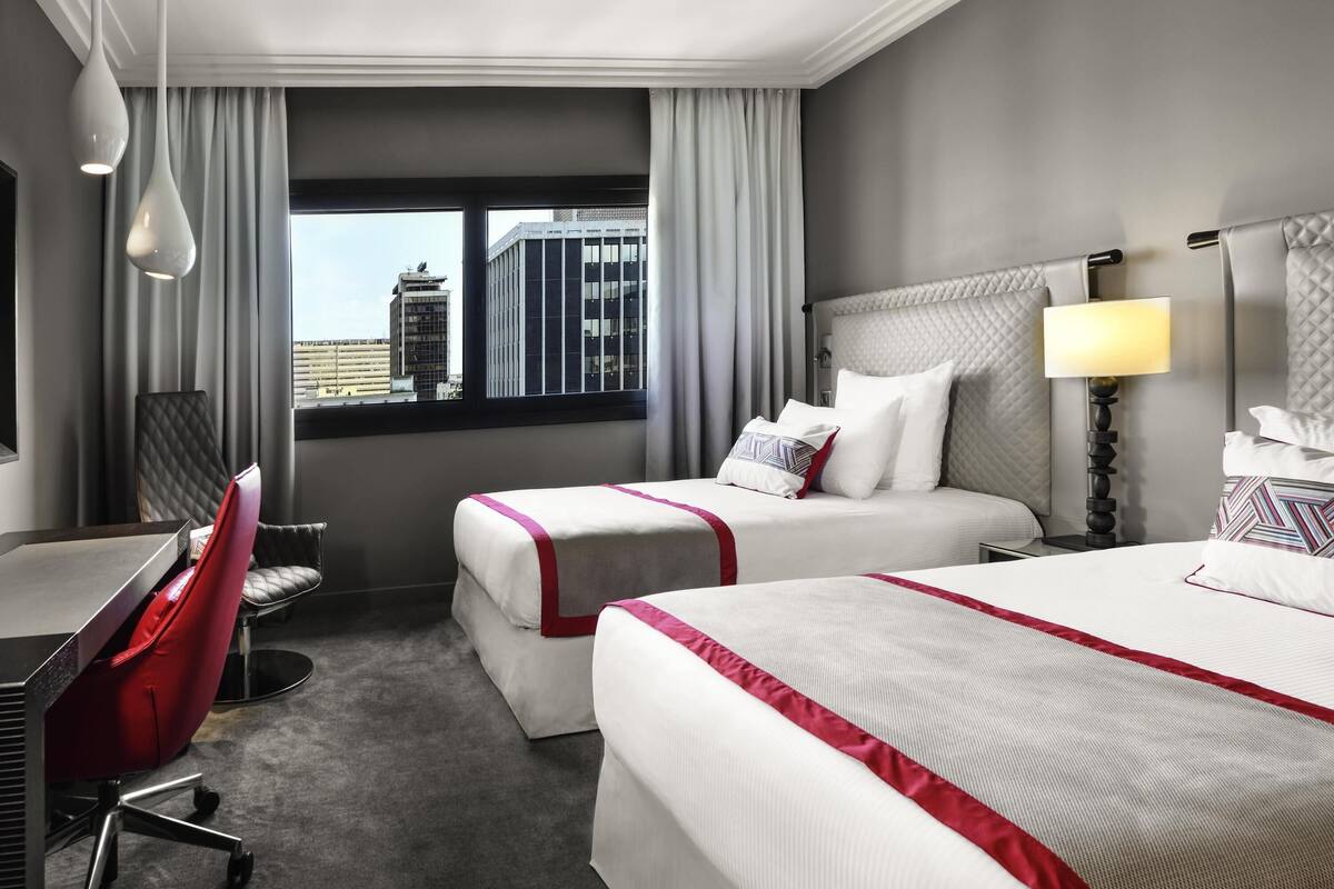 Ett hotellrum med två sängar, ett skrivbord, en stol, en lampa och utsikt över byggnader genom fönstret.