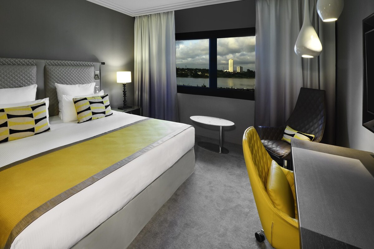 Habitación de hotel con una cama grande, un escritorio, una silla, una mesita y vistas a un paisaje urbano por la ventana.
