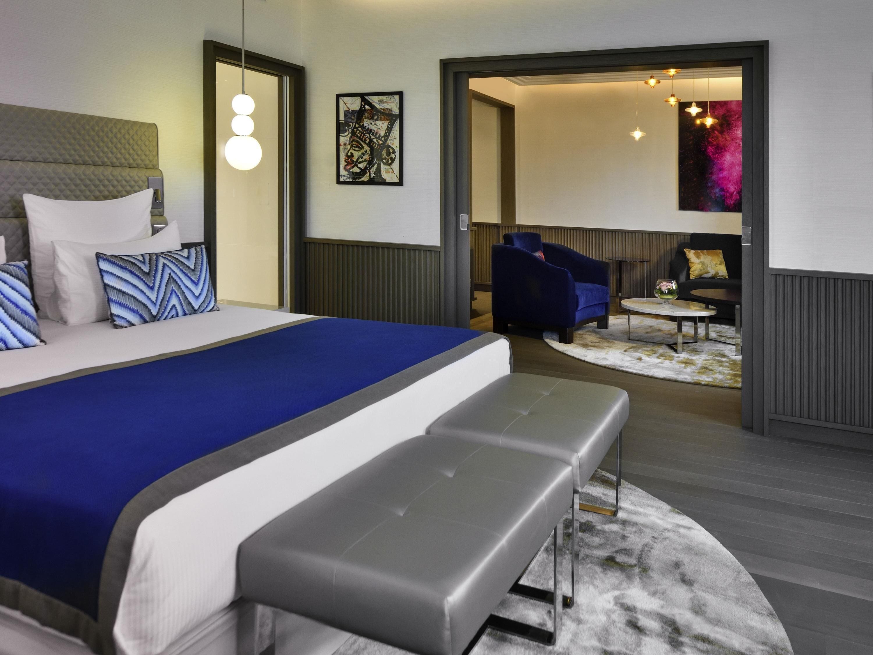 A modern hotel room with a large bed, a bench, a sofa, and a dining table.