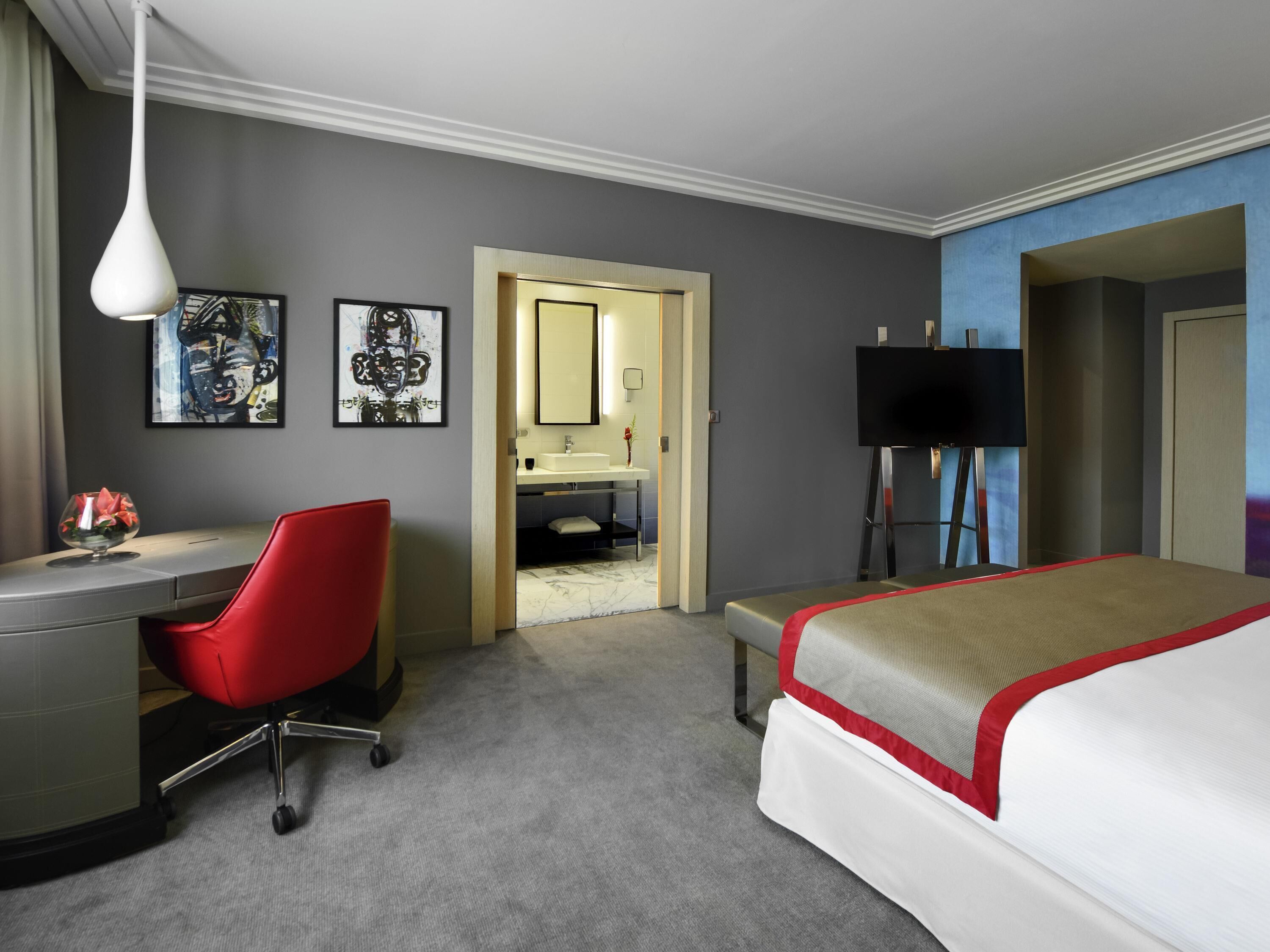 A hotel room with a desk, a red chair, a bed, a TV, and artwork on the walls.